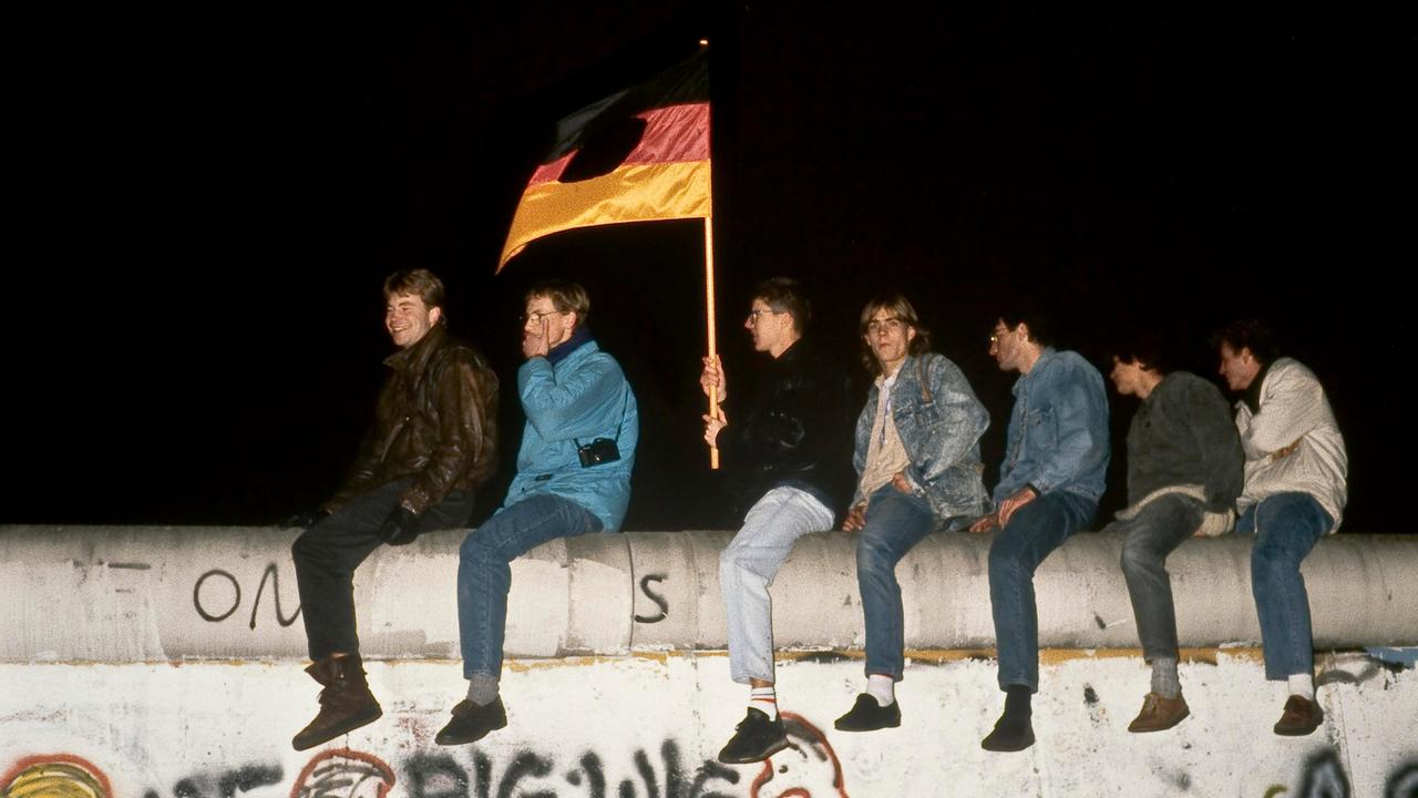 In der Nacht des 9. November 1989 sind Menschen mit einer zerschnittenen DDR-Fahne auf die Berliner Mauer am Brandenburger Tor geklettert.