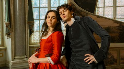 Die Schauspieler Havana Joy und Eren M. Güvercin stehen in barocken Kostümen als Geschwister Mozart vor einem aufgeklappten Cembalo.