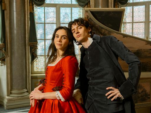 Die Schauspieler Havana Joy und Eren M. Güvercin stehen in barocken Kostümen als Geschwister Mozart vor einem aufgeklappten Cembalo.