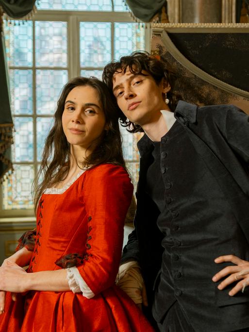 Die Schauspieler Havana Joy und Eren M. Güvercin stehen in barocken Kostümen als Geschwister Mozart vor einem aufgeklappten Cembalo.