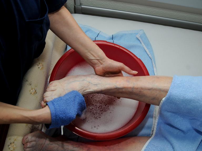 Altenpflege: Einem Patienten werden über einer Schüssel im Bett die Füsse gewaschen.