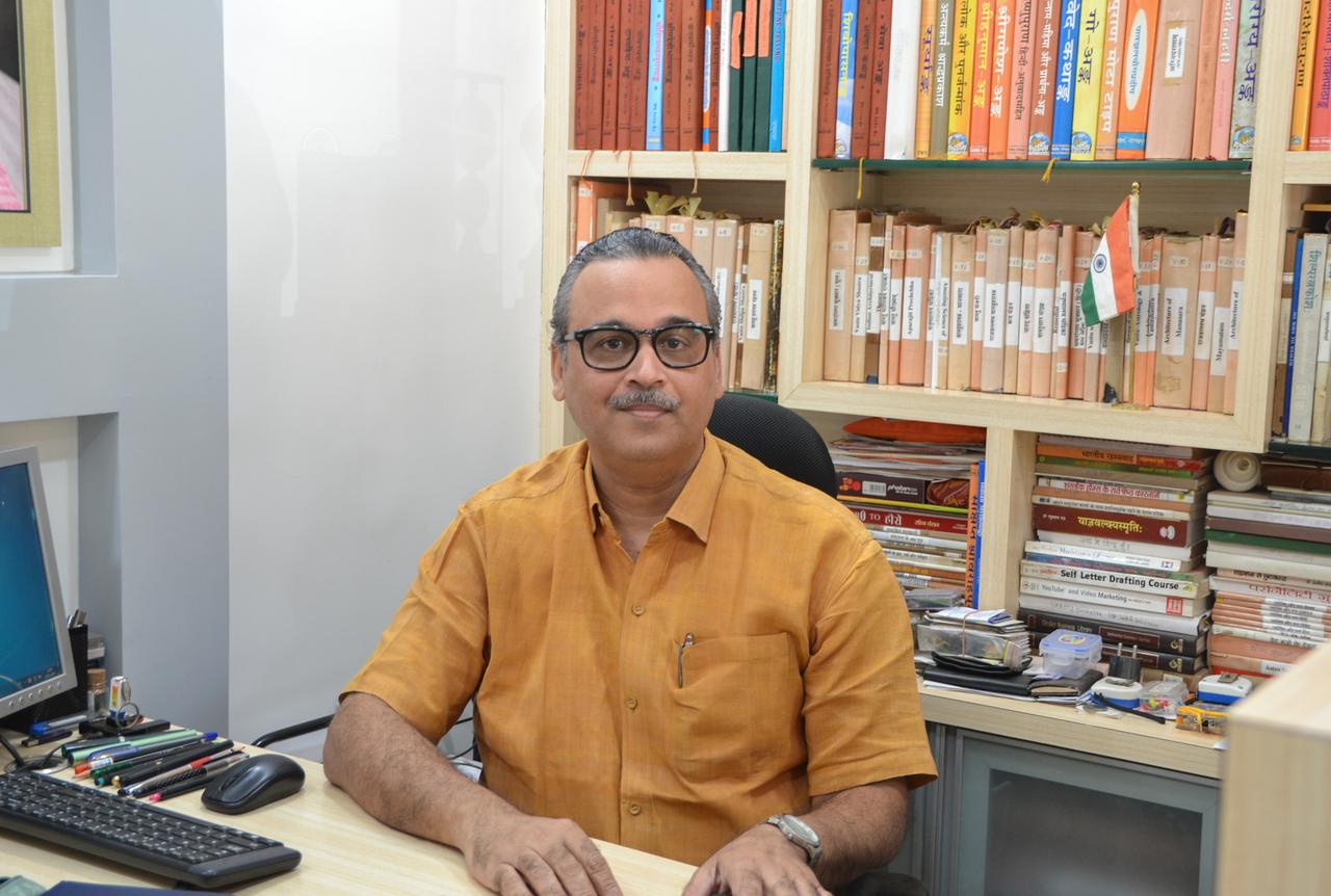 Ashok Sharma sitzt an seinem Schreibtisch, auf dem ordentlich in Reih und Glied seine Stifte liegen. Im Hintergrund ein volles Bücherregal, an dem kleine indische Flaggen angebracht sind.