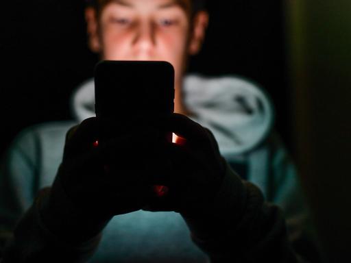 Eine unscharf abgebildete männliche Person sitzt in einem dunklen Raum und hält ein Smartphone vor sich. Das Gesicht im Hintergrund ist verschwommen, die Hände werden vom Licht des Geräts schwach beleuchtet. Eine unscharf abgebildete männliche Person sitzt in einem dunklen Raum und hält ein Smartphone vor sich. Das Gesicht im Hintergrund ist verschwommen, die Hände werden vom Licht des Geräts schwach beleuchtet.