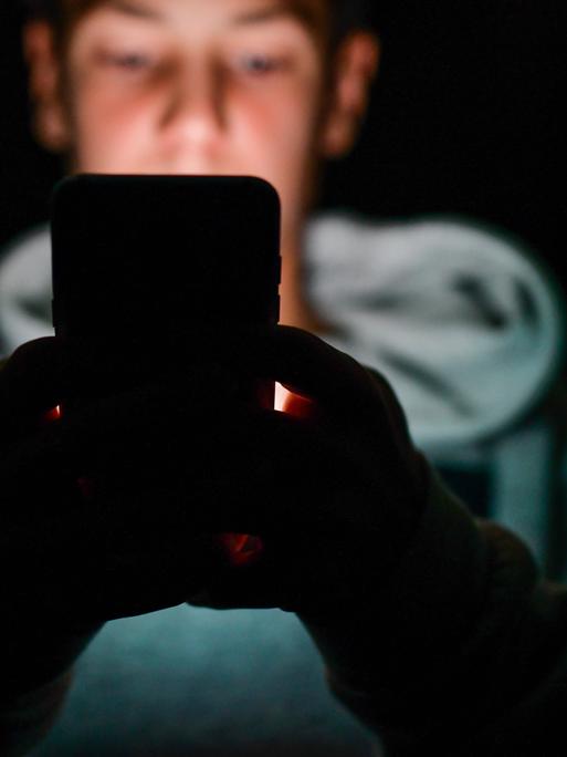 Eine unscharf abgebildete männliche Person sitzt in einem dunklen Raum und hält ein Smartphone vor sich. Das Gesicht im Hintergrund ist verschwommen, die Hände werden vom Licht des Geräts schwach beleuchtet.