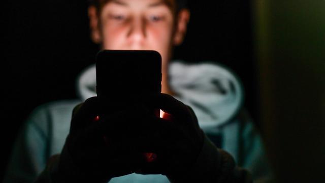 Eine unscharf abgebildete männliche Person sitzt in einem dunklen Raum und hält ein Smartphone vor sich. Das Gesicht im Hintergrund ist verschwommen, die Hände werden vom Licht des Geräts schwach beleuchtet.