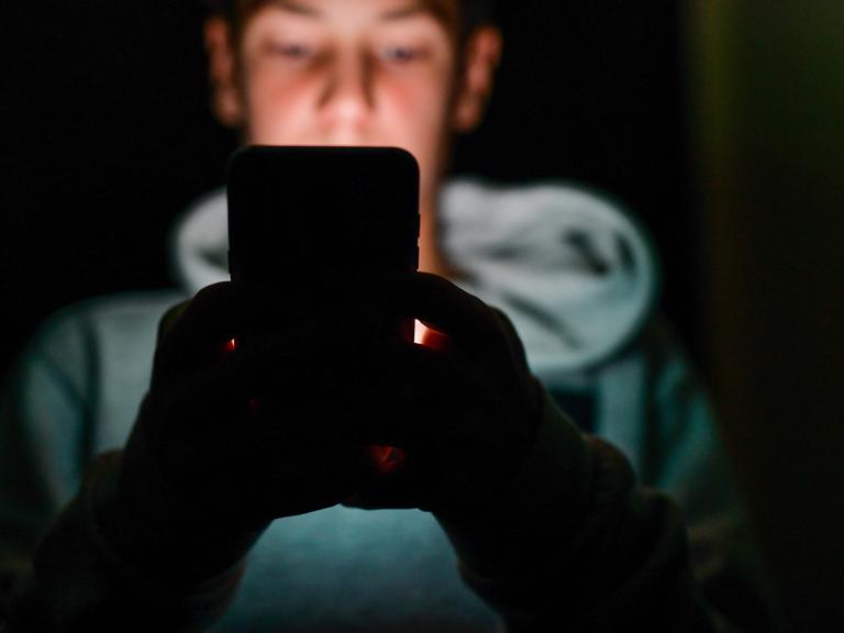 Eine unscharf abgebildete männliche Person sitzt in einem dunklen Raum und hält ein Smartphone vor sich. Das Gesicht im Hintergrund ist verschwommen, die Hände werden vom Licht des Geräts schwach beleuchtet.
