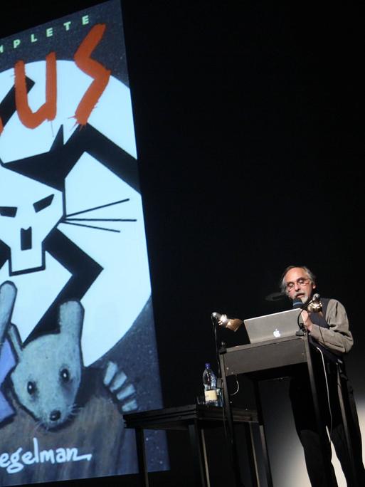 Ein an die Wand projiziertes Comiccover. Oben steht "The complete Maus". Darunter ein gemalter Katzenkopf mit Hitlerbart vor einem Hakenkreuz. Darunter zwei Comicmäuse. Daneben steht ein Mann am Pult, schaut in seinen Computer und spricht in ein Mikrofon.
