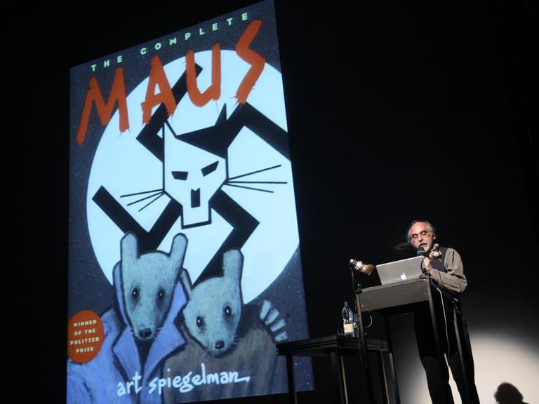 Ein an die Wand projiziertes Comiccover. Oben steht "The complete Maus". Darunter ein gemalter Katzenkopf mit Hitlerbart vor einem Hakenkreuz. Darunter zwei Comicmäuse. Daneben steht ein Mann am Pult, schaut in seinen Computer und spricht in ein Mikrofon.