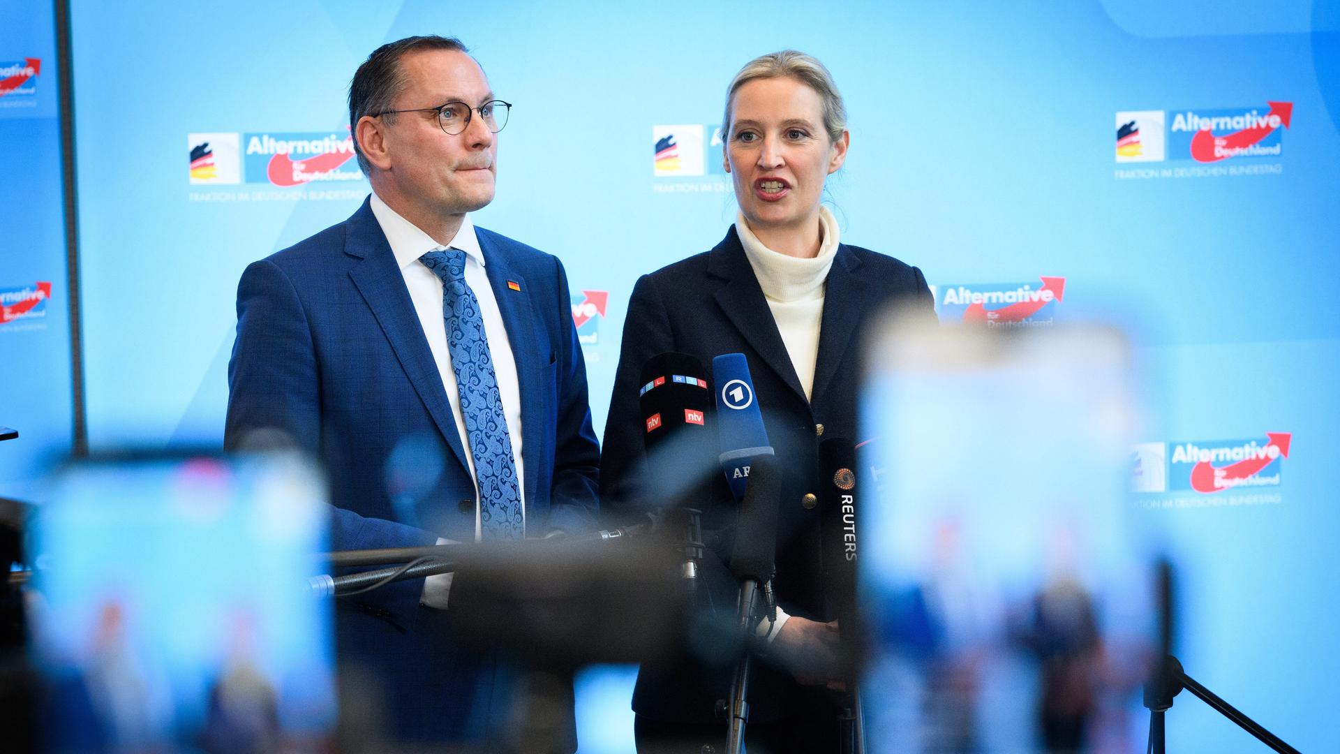 Die AfD-Politiker Tino Chrupalla und Alice Weidel stehten vor einer blauen Wand mit AfD-Logos. Vor ihnen stehen Mikrofone und Kameras.