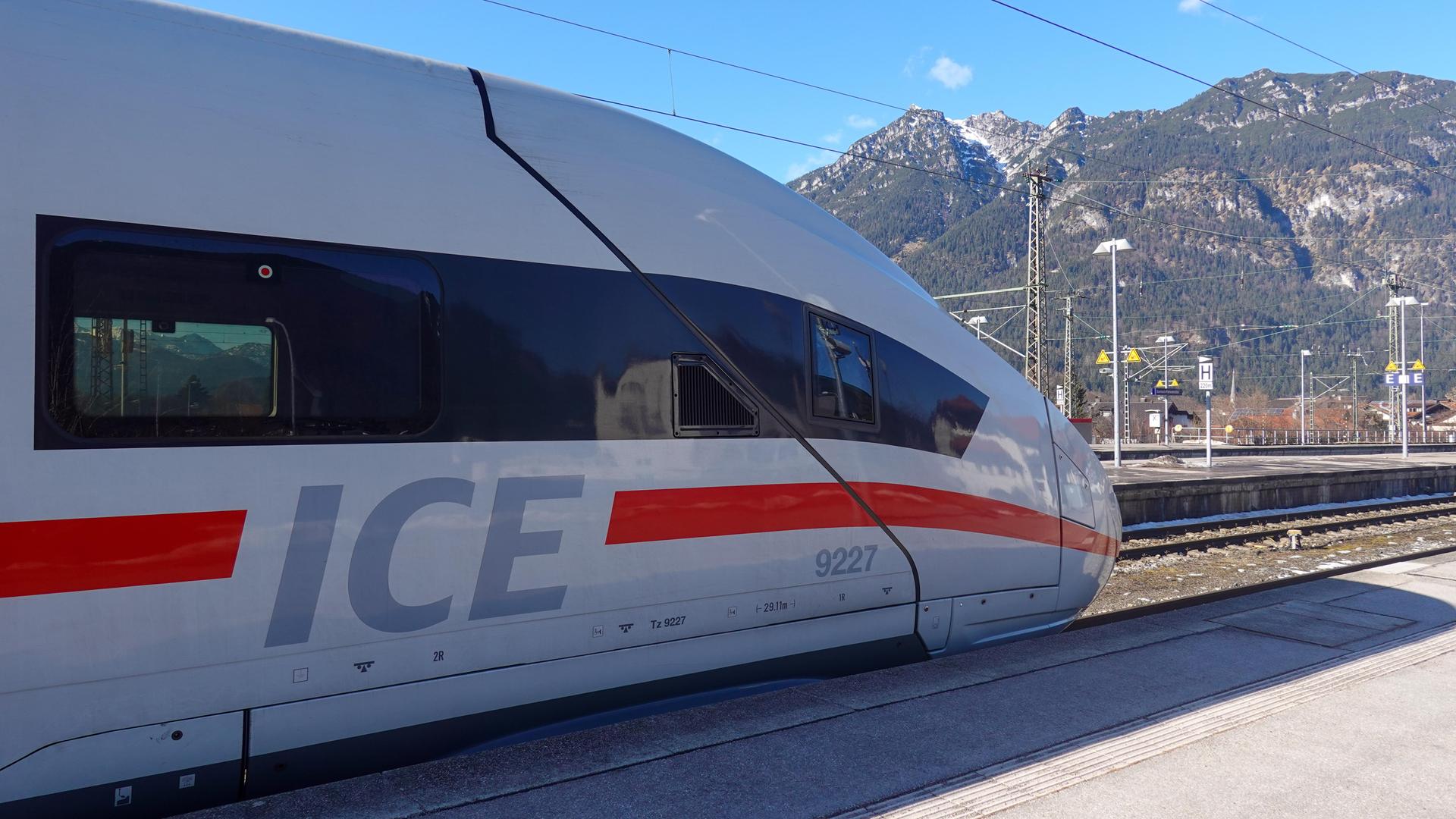 Ein Zug der Deutschen Bahn fährt auf einen Bahnsteig. Im Hintergrund sieht man hohe Berge.