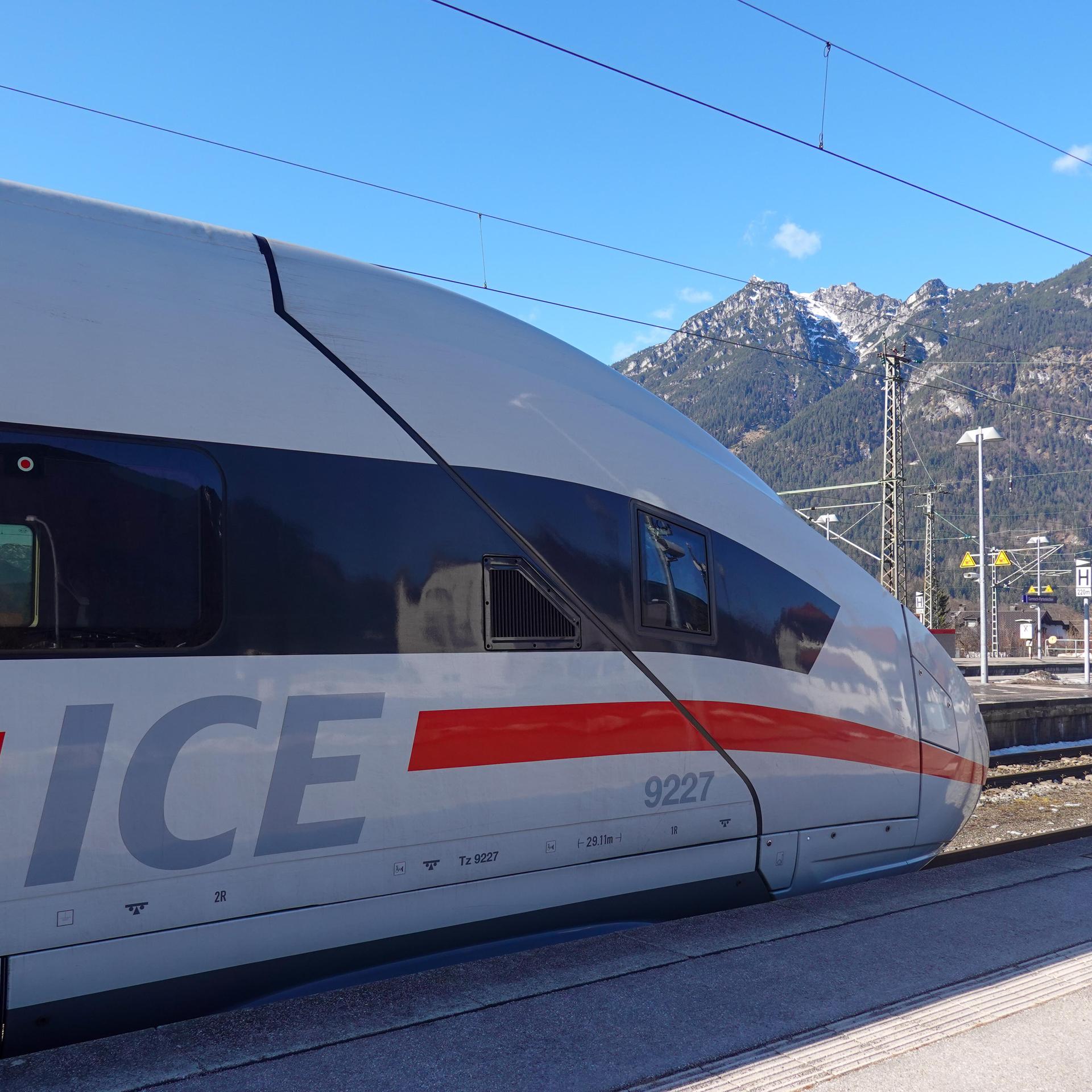 Ein Zug der Deutschen Bahn fährt auf einen Bahnsteig. Im Hintergrund sieht man hohe Berge.