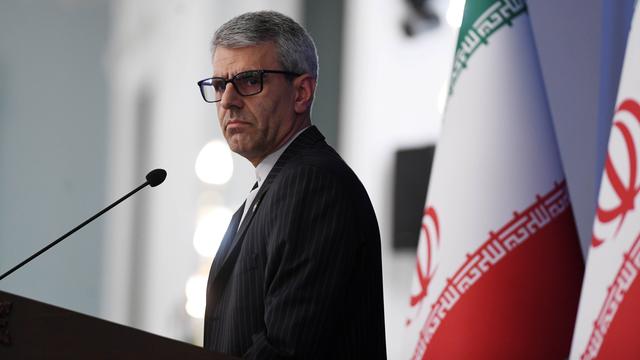 Ein Mann mit grauen Haaren und Brille schaut ernst. Er spricht in ein Mikrofon. Neben ihm sind zwei große iranische Flaggen. 