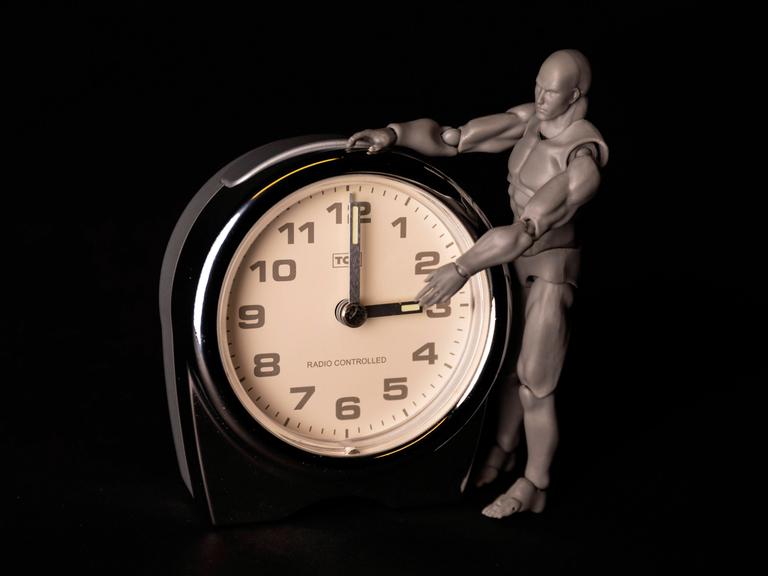 Eine kleine graue Menschenfigur steht neben dem Ziffernblatt eines Weckers; ihr linker Arm greift nach dem Zeiger, der auf drei Uhr steht.