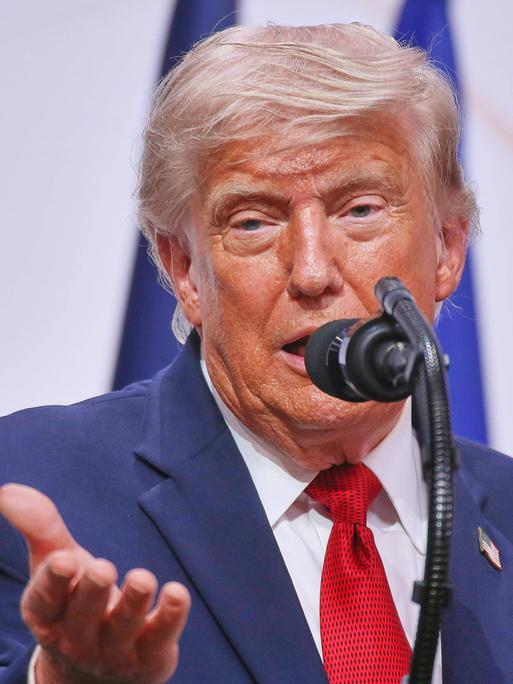 Donald Trump spricht in ein Mikrofon und hebt dabei die rechte Handfläche.