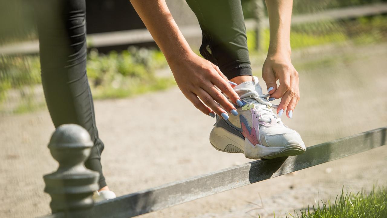 Hände binden bei Joggingschuhen die Schnürsenkel. Die Fingernägel sind lackiert. Es scheinen die Hände eine weiblich gelesenen Person zu sein.