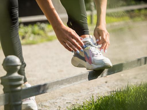 Hände binden bei Joggingschuhen die Schnürsenkel. Die Fingernägel sind lackiert. Es scheinen die Hände eine weiblich gelesenen Person zu sein.