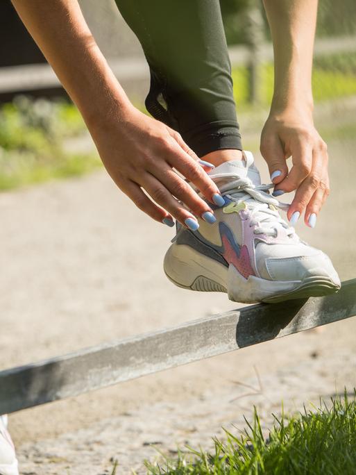 Hände binden bei Joggingschuhen die Schnürsenkel. Die Fingernägel sind lackiert. Es scheinen die Hände eine weiblich gelesenen Person zu sein.