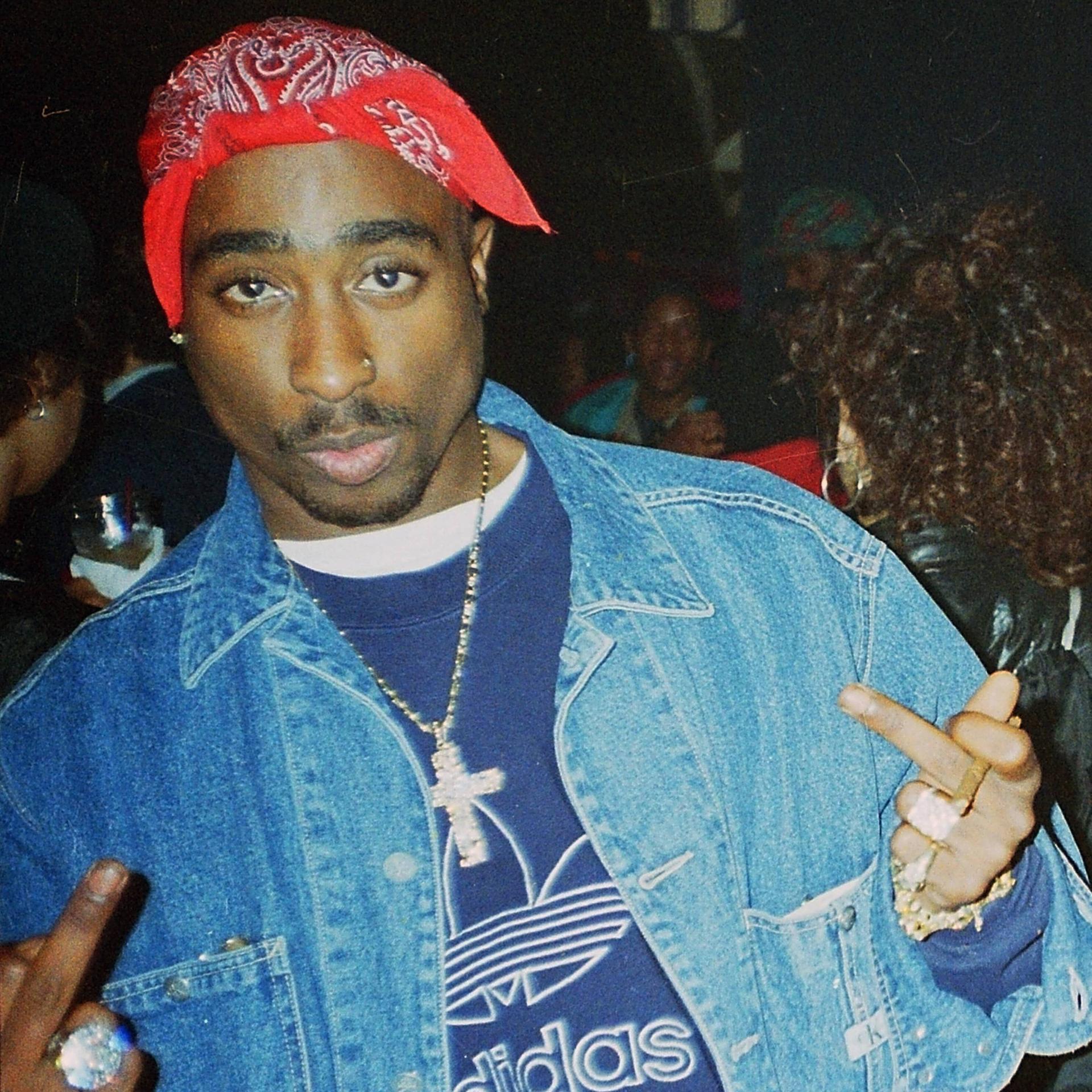 Tupac Shakur mit rotem gebundenen Tuch auf dem Kopf in den Straßen von New York, während er direkt in die Kamera blickt und die Mittelfinger nach oben streckt.