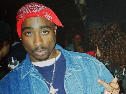 Tupac Shakur mit rotem gebundenen Tuch auf dem Kopf in den Straßen von New York, während er direkt in die Kamera blickt und die Mittelfinger nach oben streckt.
