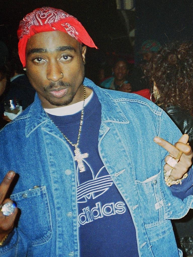 Tupac Shakur mit rotem gebundenen Tuch auf dem Kopf in den Straßen von New York, während er direkt in die Kamera blickt und die Mittelfinger nach oben streckt.