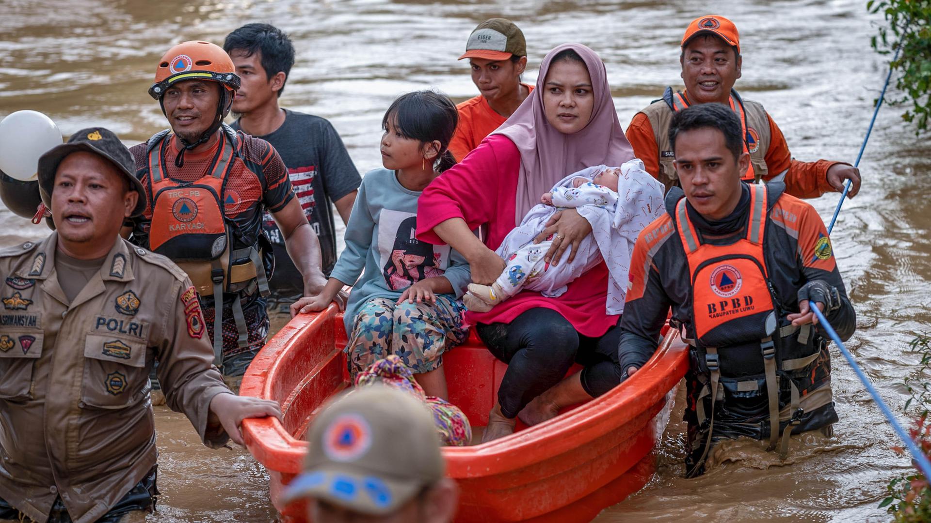 Eine Frau sitzt mit ihrem Baby und einem Mädchen in einem Rettungsboot. Sie werden von Rettern durchs Wasser geschoben.
