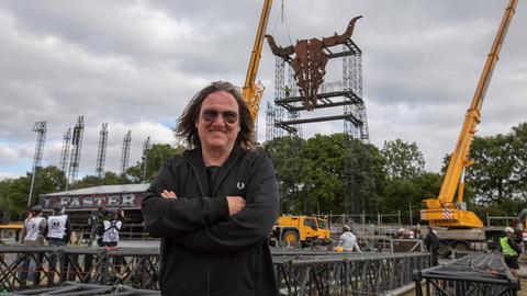 Thomas Jensen steht vor den aufwändigen Aufbauarbeiten des Festivalgeländes. Hinter ihm ist ein großer, aus rostigem Metall gestalteter Kuhschädel zu sehen, der an einer turmhohen Metallkonstruktion aufgehängt ist.