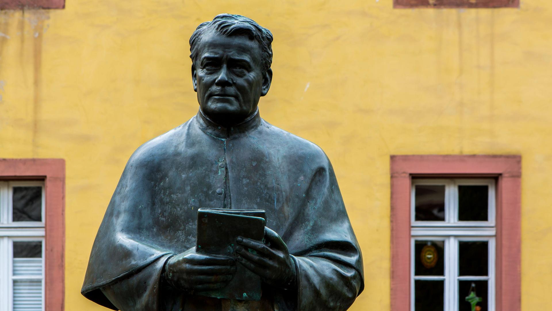 Eine Bronzestatue steht auf einem Innnenhof vor einer gelben Wand mit rot eingefassten Fenstern.