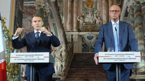 Die beiden stehen nebeneinander an Rednerpulten vor einem Eingang. Macron sagt etwas und ballt dazu beide Fäuste. Neben den beiden Fahnen der EU, Frankreichs und Deutschlands. 