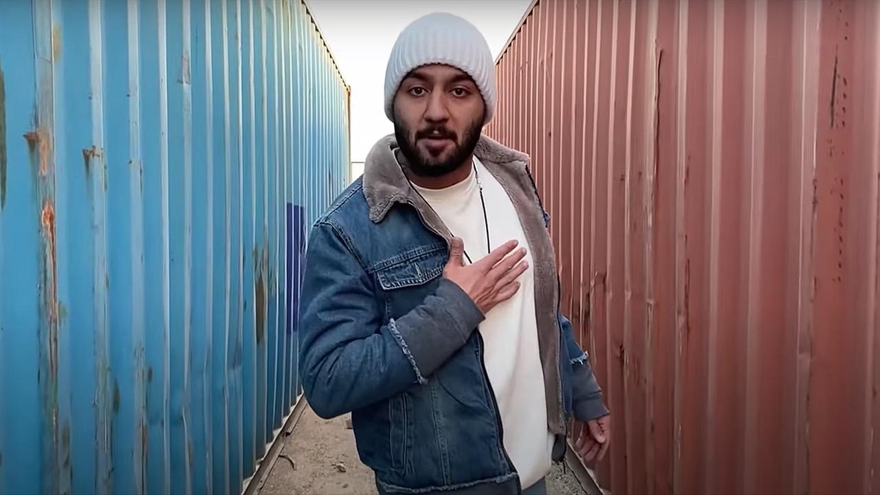 Das Bild zeigt den iranischen Rapper Toomaj Salehi. Er steht mit Wollmütze und Jeansjacke zwischen zwei Containern und fasst sich ans Herz.