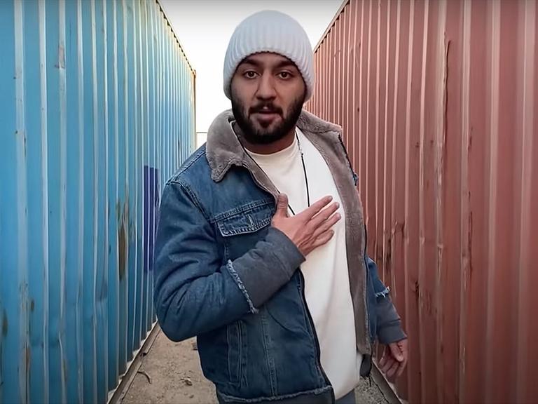 Der iranische Rapper Toomaj Salehi steht mit Wollmütze und Jeansjacke zwischen zwei Containern und fasst sich ans Herz.