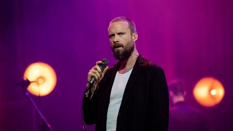Father John Misty steht in dunkler Kleidung auf einer Bühne mit gelblichen Scheinwerfern, die wie große Glühbirnen aussehen.