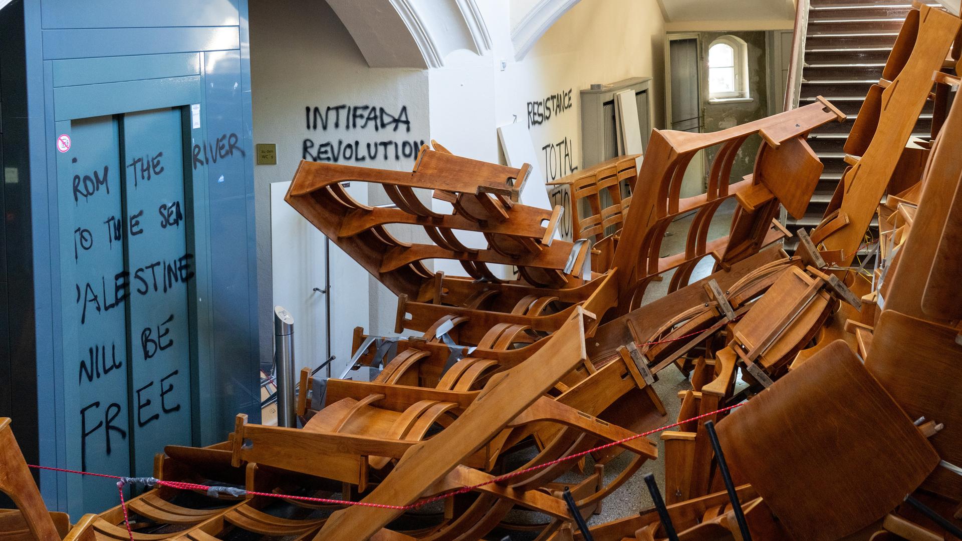 In einem Treppenflur der Humboldt-Universität zu Berlin stapeln sich herausgerissene Holzstuhlreihen vor dem Eingang eines Vorlesungssaals. Die Wände sind mit propalästinensischen und hamas-nahen Parolen beschmiert