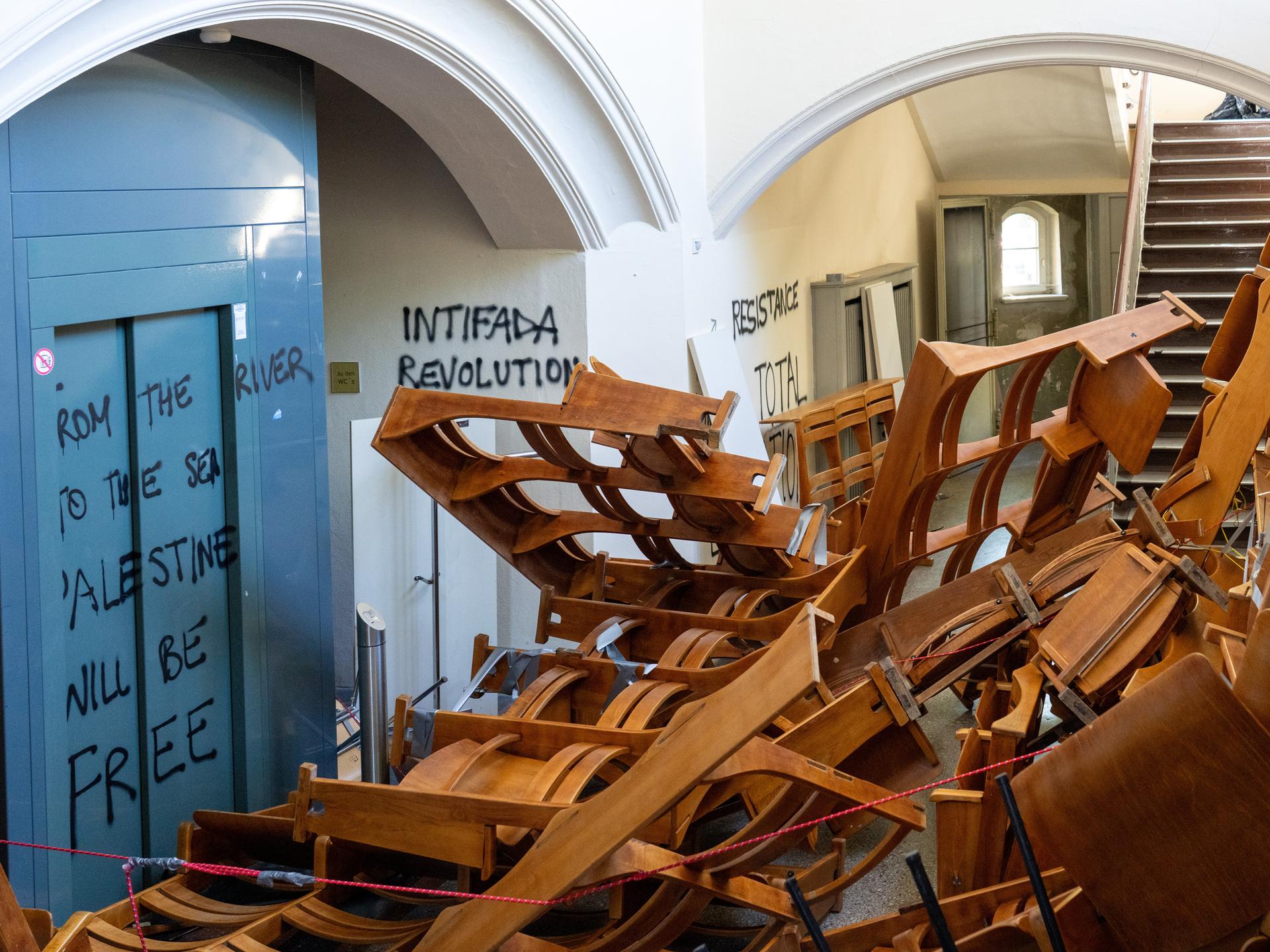 In einem Treppenflur der Humboldt-Universität zu Berlin stapeln sich herausgerissene Holzstuhlreihen vor dem Eingang eines Vorlesungssaals. Die Wände sind mit propalästinensischen und hamas-nahen Parolen beschmiert