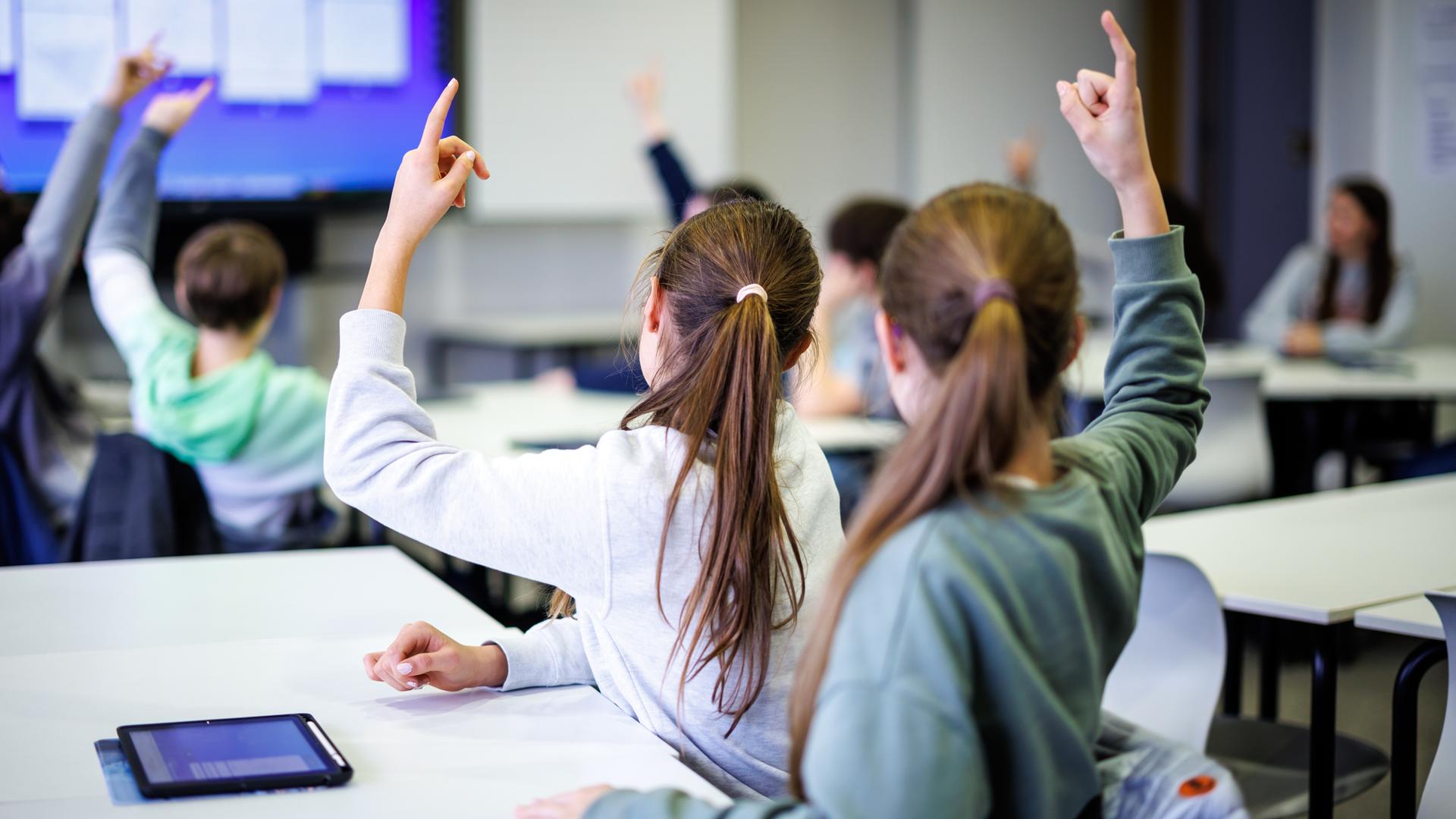 Schülerinnen und Schüler sitzen in einem Klassenraum. Einige melden sich. Auf einem Tisch liegt ein Tablet. Schülerinnen und Schüler sitzen in einem Klassenraum. Einige melden sich. Auf einem Tisch liegt ein Tablet.