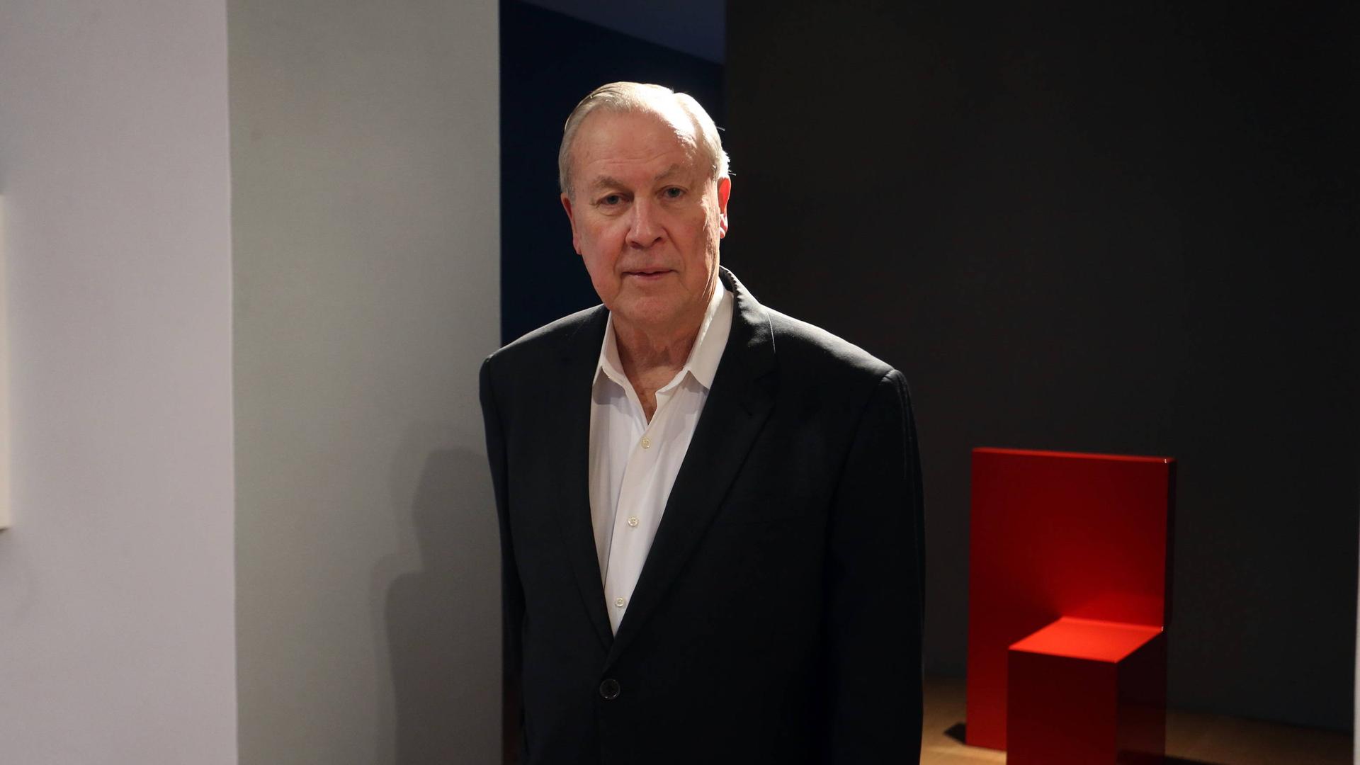 Robert Wilson steht im schwarzen Anzug vor einem Ausstellungsraum, in dem ein schlichtes rotes Möbelobjekt zu sehen ist. Robert Wilson steht im schwarzen Anzug vor einem Ausstellungsraum, in dem ein schlichtes rotes Möbelobjekt zu sehen ist.