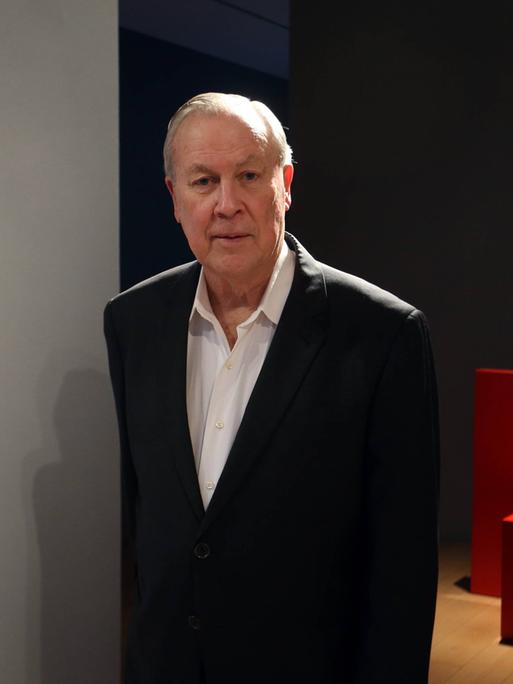 Robert Wilson steht im schwarzen Anzug vor einem Ausstellungsraum, in dem ein schlichtes rotes Möbelobjekt zu sehen ist.