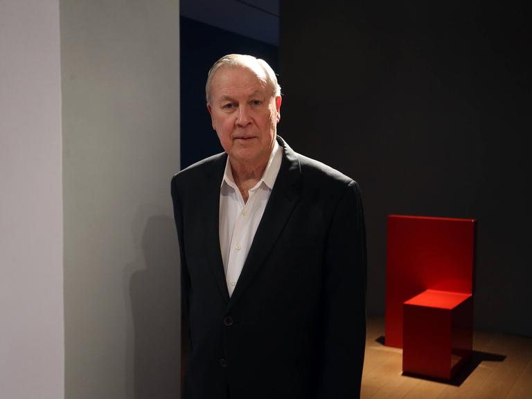 Robert Wilson steht im schwarzen Anzug vor einem Ausstellungsraum, in dem ein schlichtes rotes Möbelobjekt zu sehen ist.