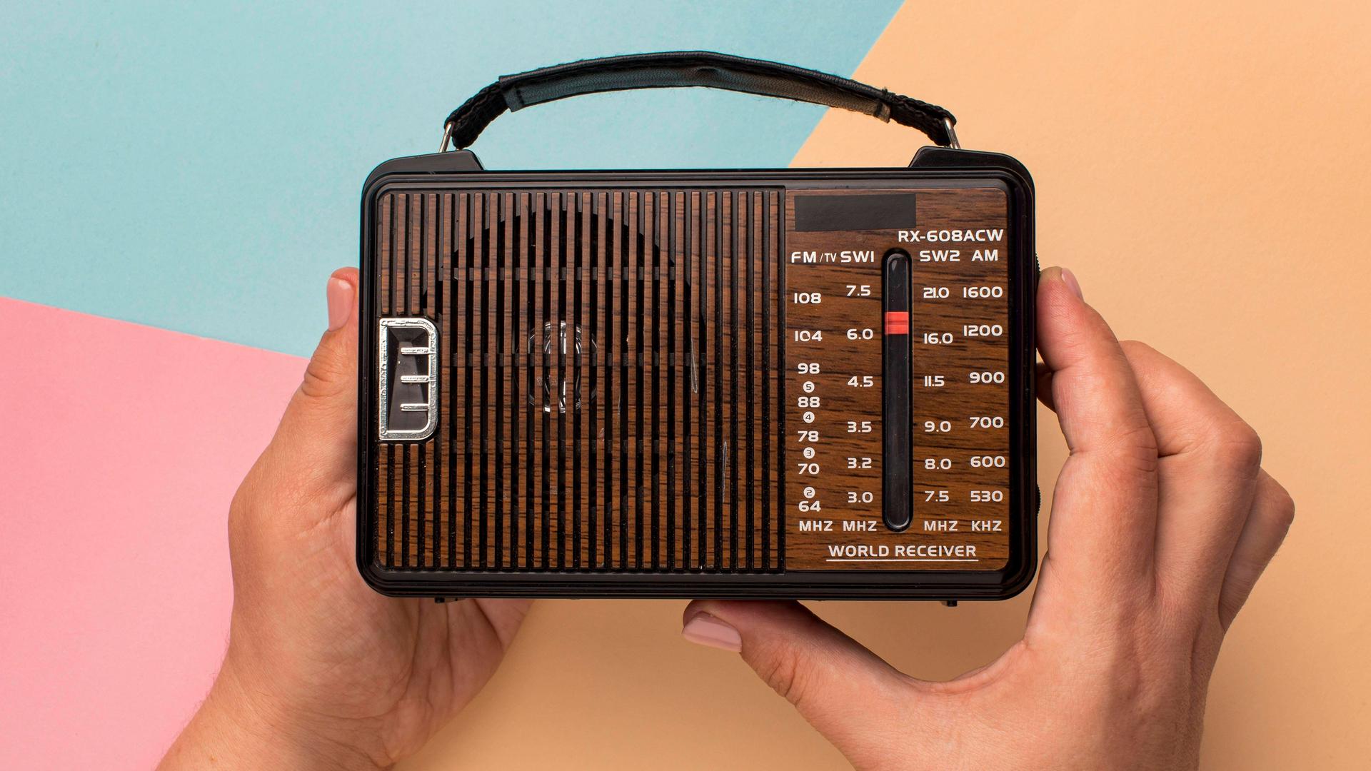 Hände halten ein kleines Retro-Radio vor einem farbigem Hintergrund aus geometrischen Formen. 