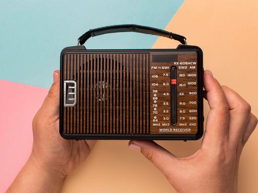 Hände halten ein kleines Retro-Radio vor einem farbigem Hintergrund aus geometrischen Formen. 