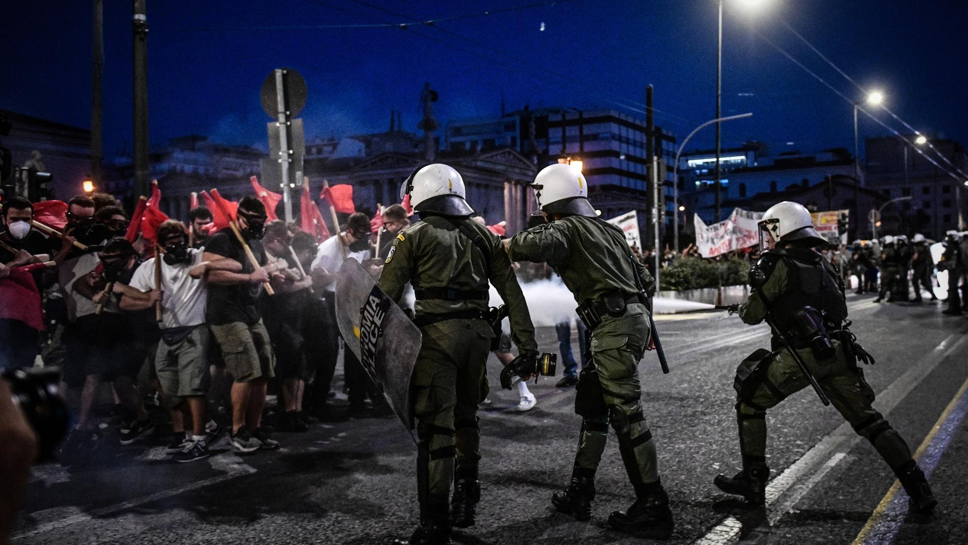 Demonstrierende mit roten Fahnen und Polizisten mit Helmen und Schutzschilden treffen auf einer nächtlichen Straße aufeinander.