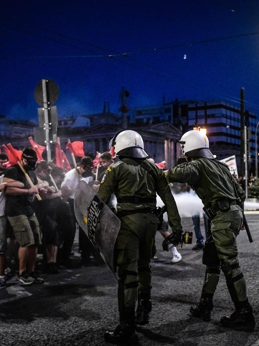 Demonstrierende mit roten Fahnen und Polizisten mit Helmen und Schutzschilden treffen auf einer nächtlichen Straße aufeinander.