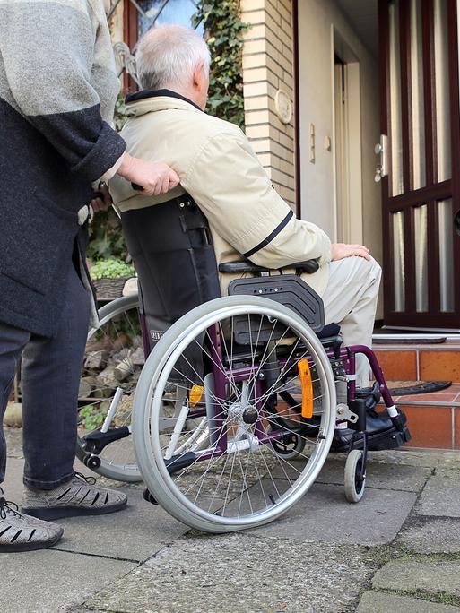 Ein älterer Mann im Rollstuhl wird am von seiner Frau vor die Haustür geschoben. 