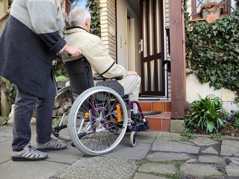 Ein älterer Mann im Rollstuhl wird am von seiner Frau vor die Haustür geschoben. 