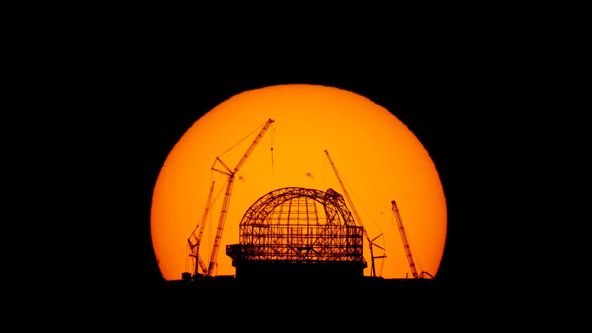 Die Aufnahme zeigt, wie ein großer orangefarbener Sonnenball hinter der Baustelle von Europas neuem Großteleskop ELT in Chile versinkt.