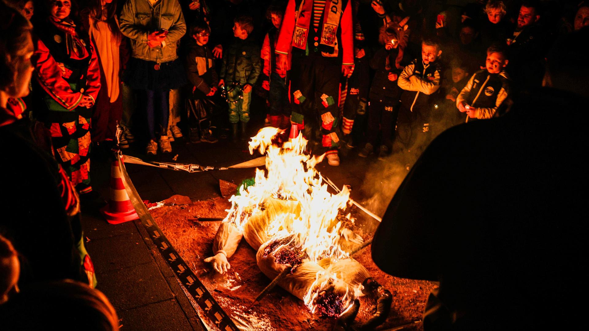Es ist dunkel. Der Nubbel brennt auf einem durch Band abgesperrten kleinen Podest. Um ihn herum werden Erwachsene und Kinder von den Flammen rot beleuchtet. Es ist dunkel. Der Nubbel brennt auf einem durch Band abgesperrten kleinen Podest. Um ihn herum werden Erwachsene und Kinder von den Flammen rot beleuchtet.