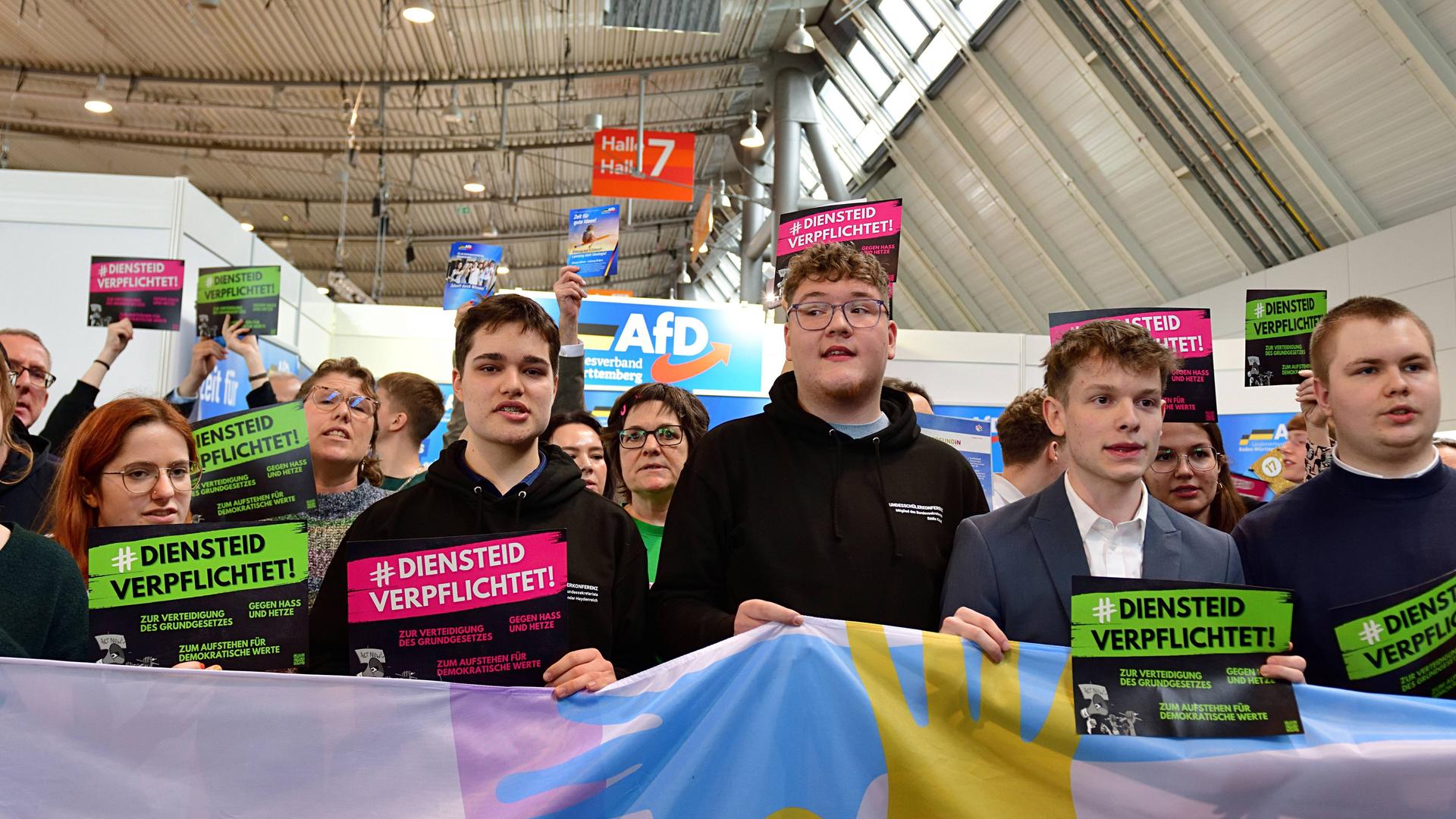 Mehrere Frauen und Männer stehen in einer messehalle. Sie tragen Schilder mit der Aufschrift "Diensteid verpflichtet". Im Hintergrund der AfD-Stand. 