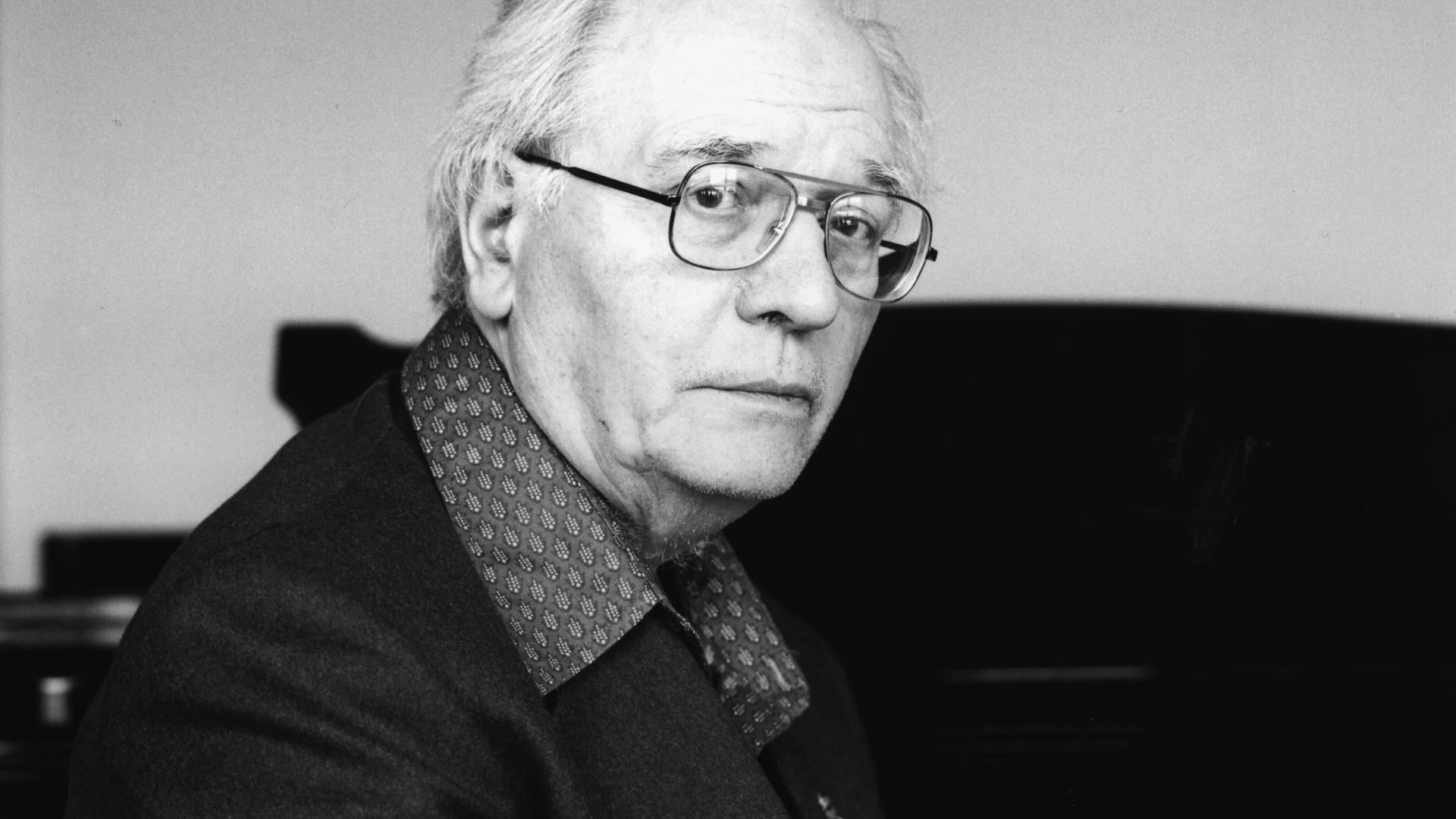 Olivier Messiaen blickt ernst durch seine Brille direkt in die Kamera, während er vor einem Flügel sitzt.