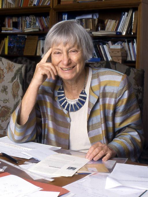 Dorothee Sölle, eine ältere Frau in einer blau-golden gestreiften Jacke, sitzt lächelnd an ihrem Schreibtisch, vor sich verschiedene Schriftstücke ausgebreitet.