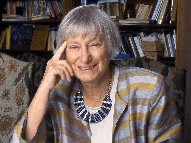 Dorothee Sölle, eine ältere Frau in einer blau-golden gestreiften Jacke, sitzt lächelnd an ihrem Schreibtisch, vor sich verschiedene Schriftstücke ausgebreitet.