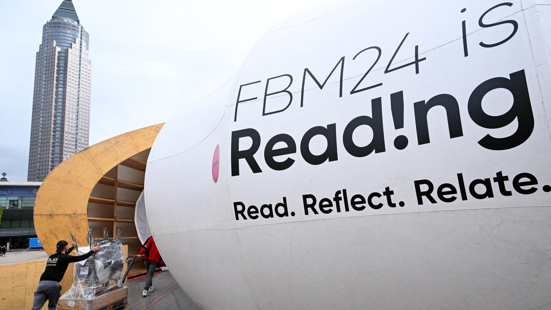 Im Hintergrund ein Tower, im Vordergrund schiebt ein Arbeiter einen Wagen in den Eingang. Daneben ein großes Schild zur Frankfurter Buchmesse, darauf steht FBM24 ist Read!ng Read. Reflect. Relate.